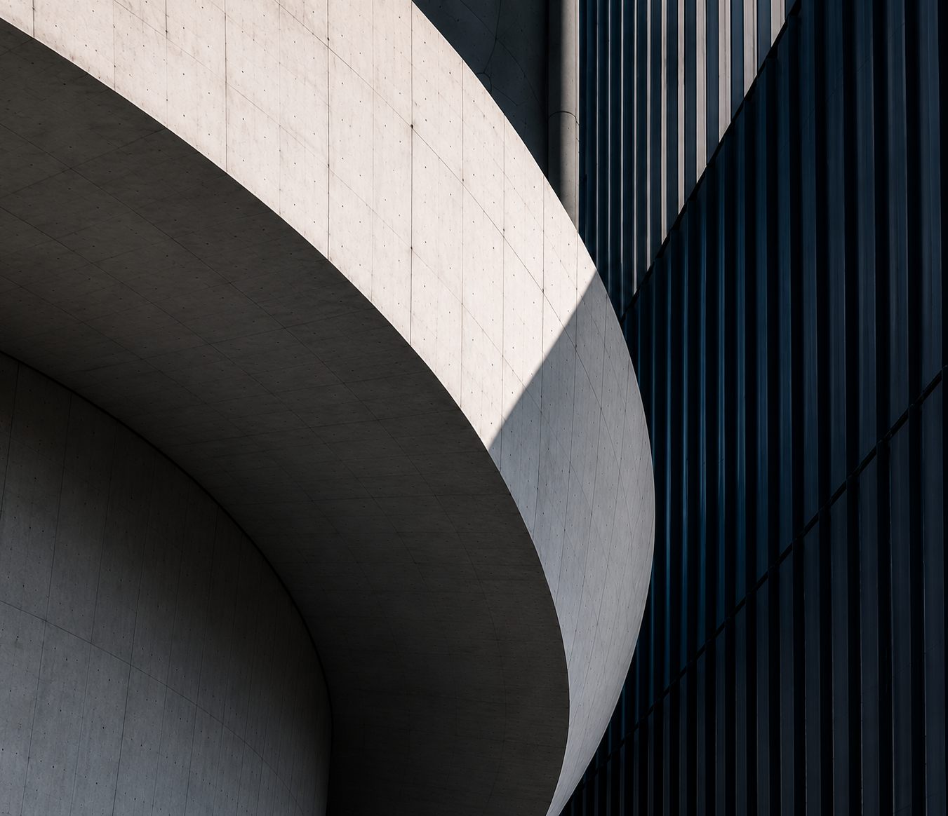 Abstract architectural detail: a curved concrete façade in hard sunlight meeting a dark ribbed wall